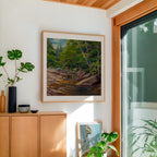 Framed landscape painting on a wall with wooden cabinet and plants in a room.