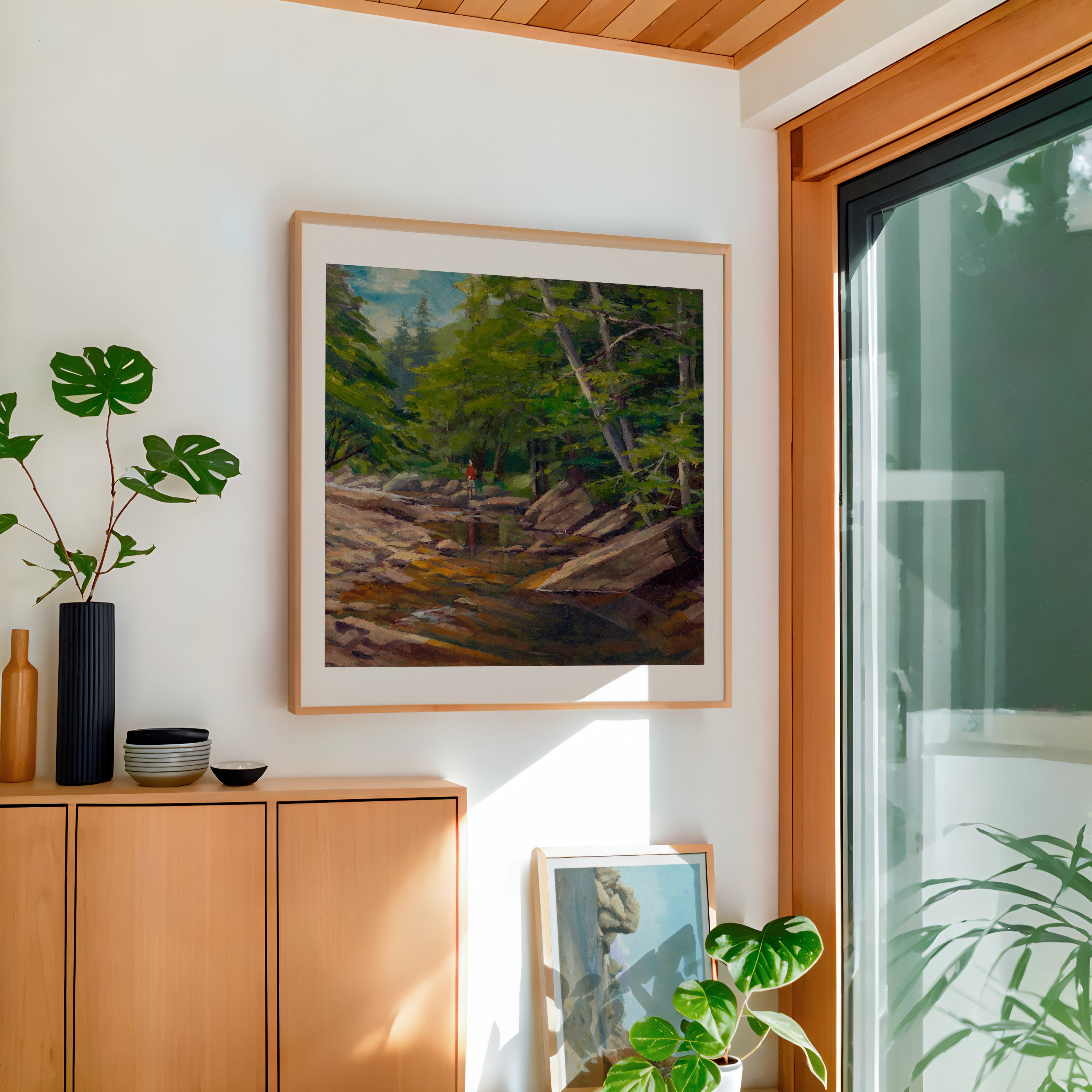 Framed landscape painting on a wall with wooden cabinet and plants in a room.