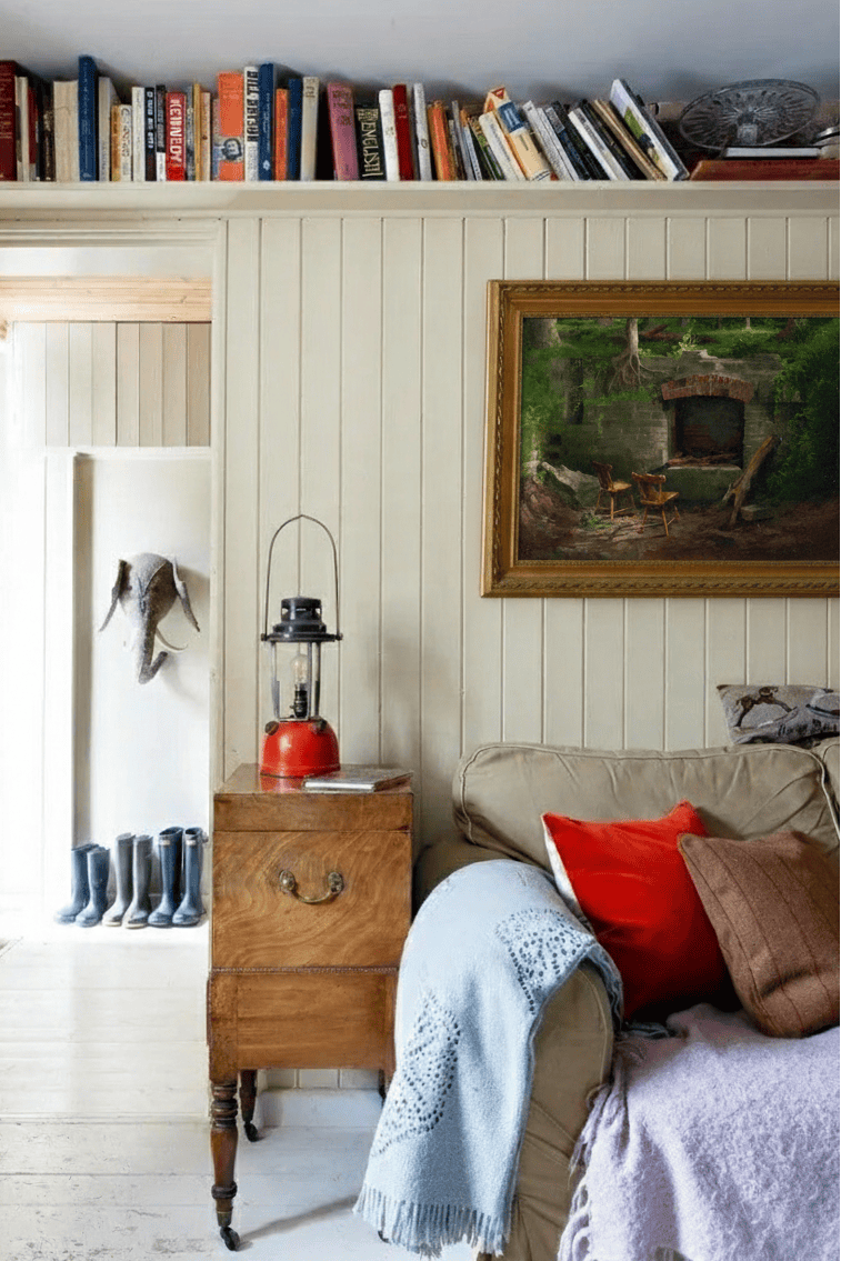 Cozy living room featuring an oil painting on the wall from artist Leo Stafsnes. The image shows a bookshelf above the couch, red oil lamp, and sofa with a matching red pillow.