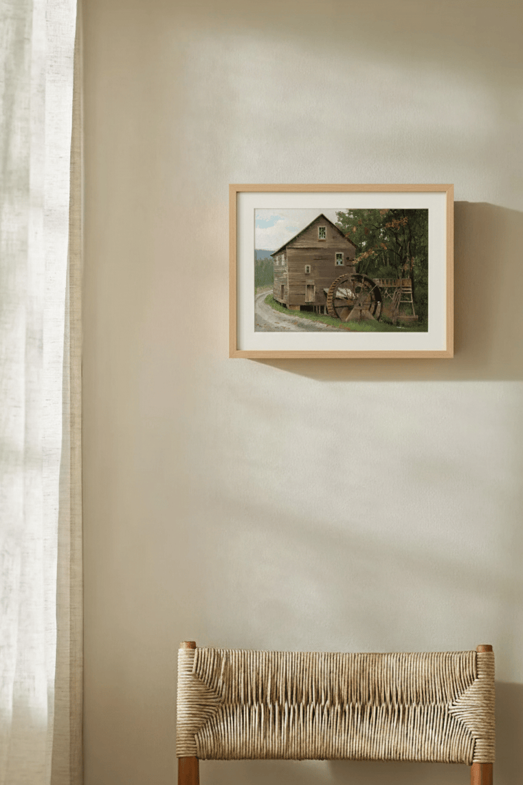Framed artwork of a water wheel by Leo Stafsnes on a wall next to a wicker chair.
