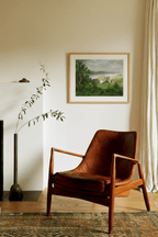 Brown leather chair in a room with a framed print by Leo Stafsnes on the wall and a curtain.